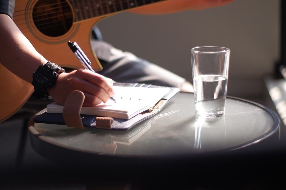 Person writing in a notebook beside an acoustic guitar and water glass